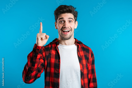 Obraz na płótnie Portrait of young thinking pondering man in red having idea moment pointing finger up on blue studio background