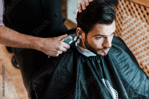 barber holding trimmer while styling hair of handsome man in barbershop