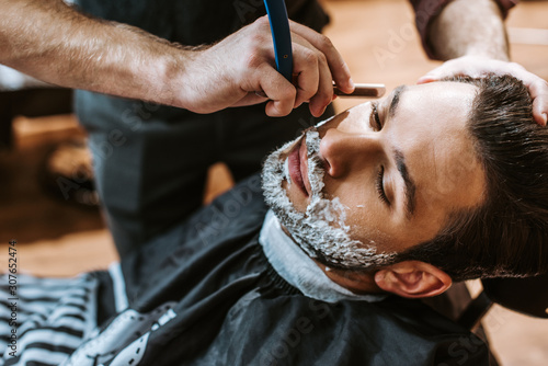 Fotografi barber shaving bearded man with shaving cream on face