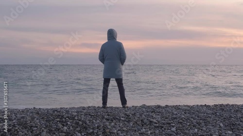 A man in a hooded jacket in cold weather stands on the beach and looks at the sea at sunset. Rear view. The zoom camera.