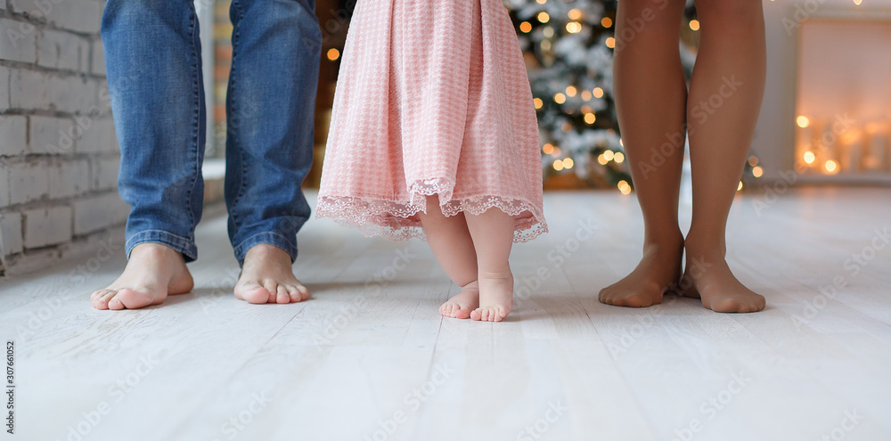 Parts of the body. Feet of dad, mom and baby. The baby takes the first steps with the help of parents, close up. Parents and their little daughter stand bare feet on a white floor against the backgrou