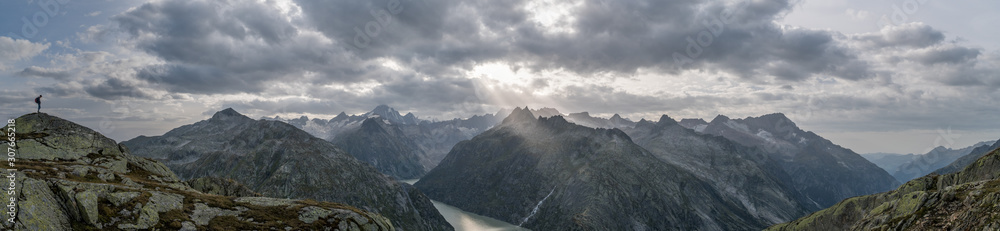 Panoramic view to the Grimsel lake in Swiss Alps with dramatic sun rays breaking through dark clouds with a boy standing on a rock