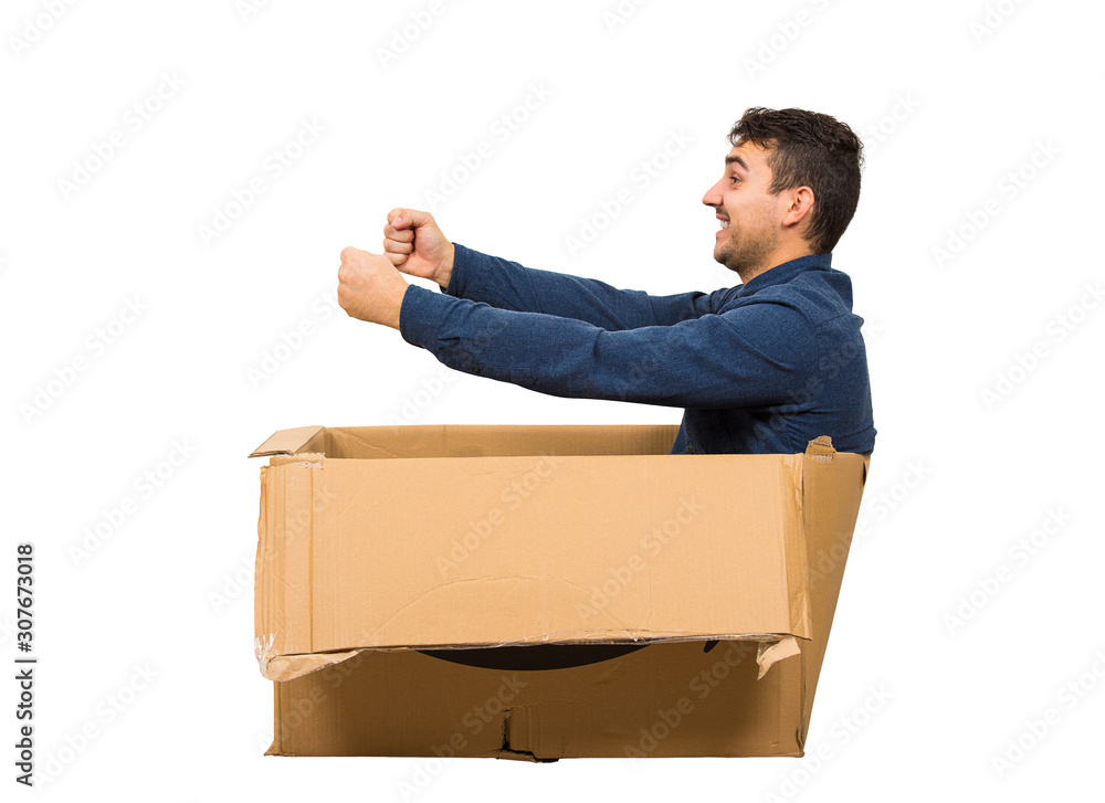 Full length side view of childish man sitting inside a cardboard box  pretending to drive a new car isolated over white background. Joyful guy  dreaming of buying a personal vehicle. Stock Photo