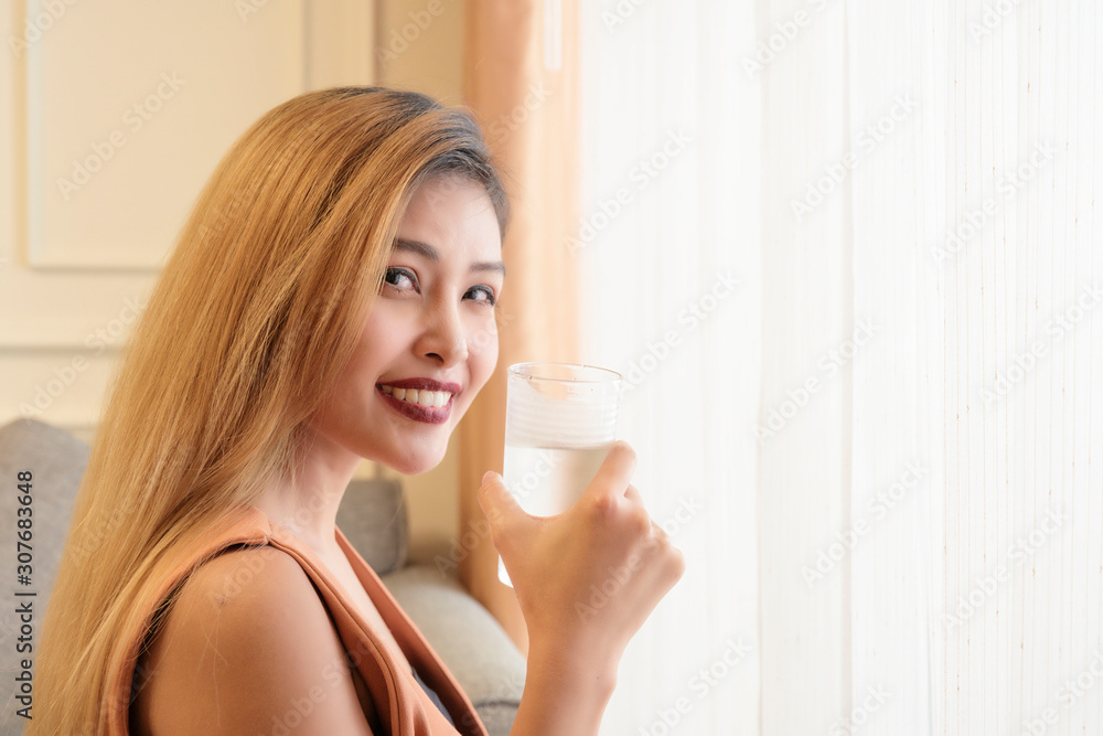 Young asian woman with gold long hair color smiling with cool glass of water in hand in room background Young asian woman with gold long hair color smiling with cool glass of water in hand in room background