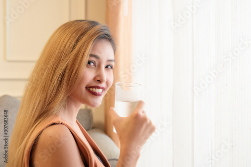 Young asian woman with gold long hair color smiling with cool glass of water in hand in room background