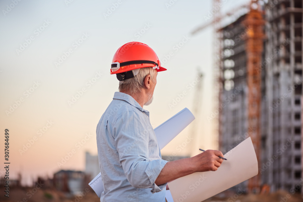 Constructor engineer with a gray beard in a constractor helmet with drawings on the background of construction Constructor engineer with a gray beard in a constractor helmet with drawings on the background of construction