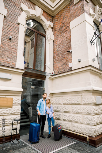 Picture of young couple entering hotel. Young man and woman standing near the old building city hotel, outdoors, holding suitcases
