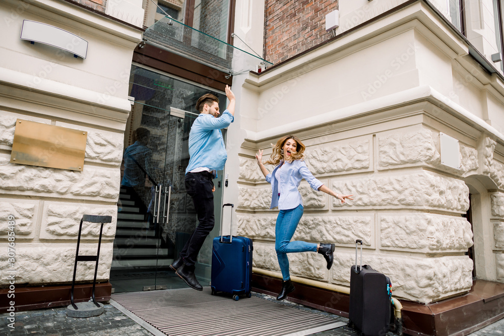 Young cheerful couple with suitcases jumping and giving five, having fun, before arriving to the hotel. Couple having fun outdoors, near the old city building