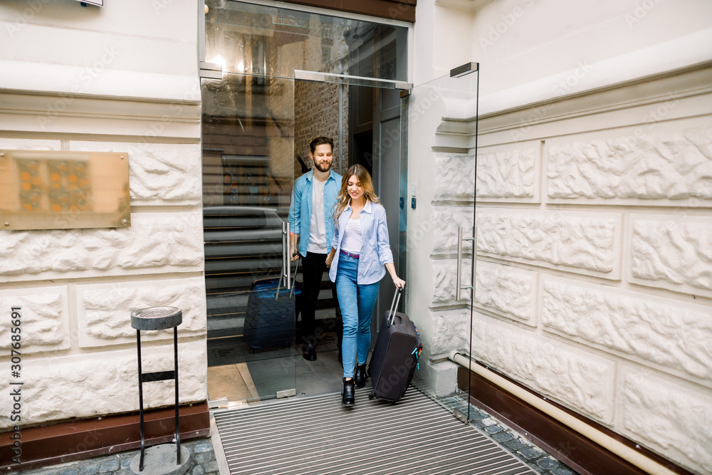 Pretty woman and handsome man holding hands are leaving a hotel with their suitcases