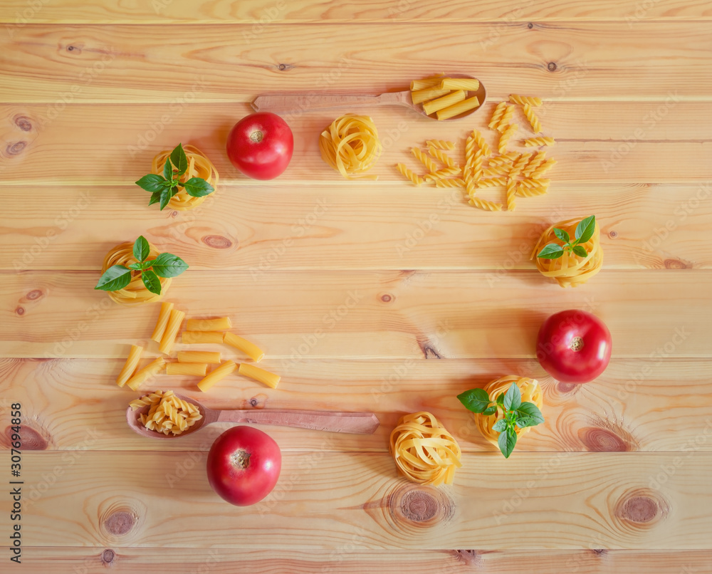 Frame made of different kinds of raw pasta, tomatoes and green basil leaves on wooden background. Top view, copy space. Frame made of different kinds of raw pasta, tomatoes and green basil leaves on wooden background. Top view, copy space.