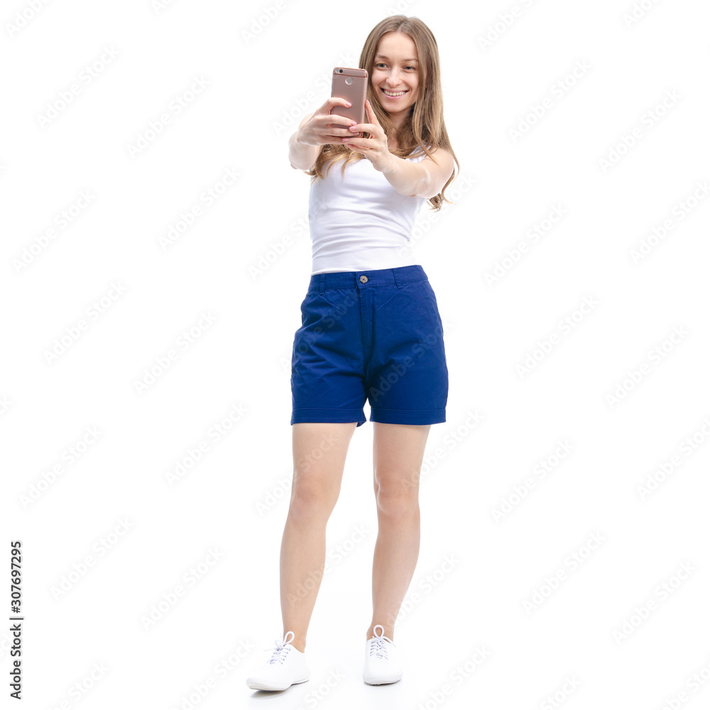 Woman in blue shorts and white shirt standing, smiling, making selfie on smartphone on white background isolation Woman in blue shorts and white shirt standing, smiling, making selfie on smartphone on white background isolation