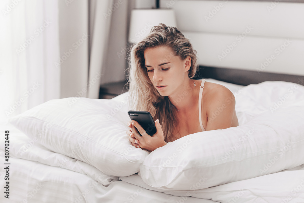 Relaxed slim young woman reads social networks in a smartphone while lying on a bed in morning. Charming blonde lady will check mail before work day. Business, communication and friends concept Relaxed slim young woman reads social networks in a smartphone while lying on a bed in morning. Charming blonde lady will check mail before work day. Business, communication and friends concept