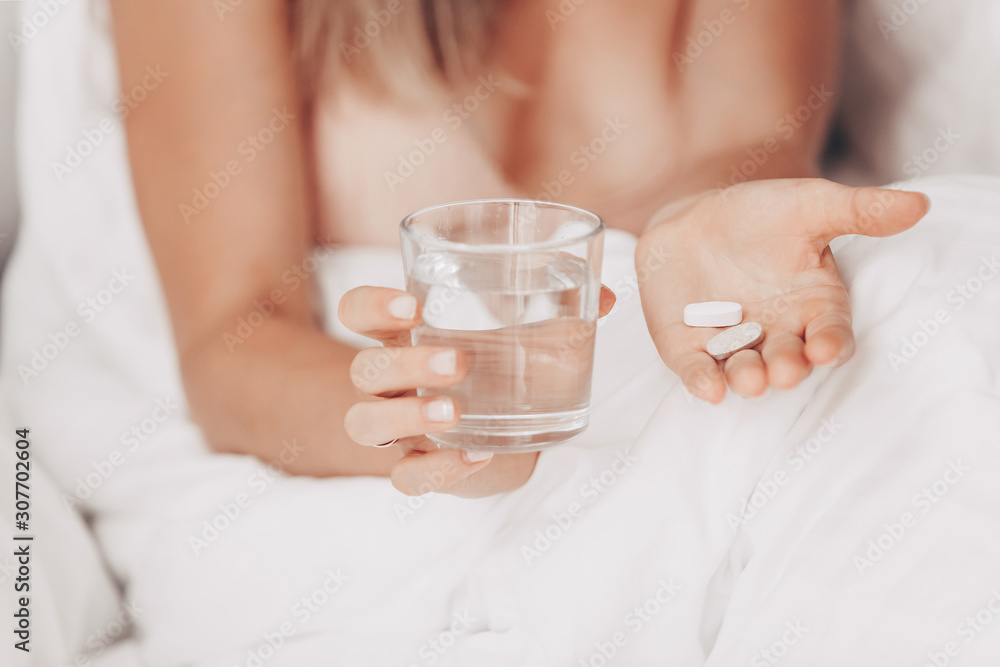 Hands of a young slim healthy caucasian girl sitting in a morning bed hold vitamins and a glass of water. Young healthy woman is drinking cold medicine. Treatment Prevention Wellness Concept Hands of a young slim healthy caucasian girl sitting in a morning bed hold vitamins and a glass of water. Young healthy woman is drinking cold medicine. Treatment Prevention Wellness Concept