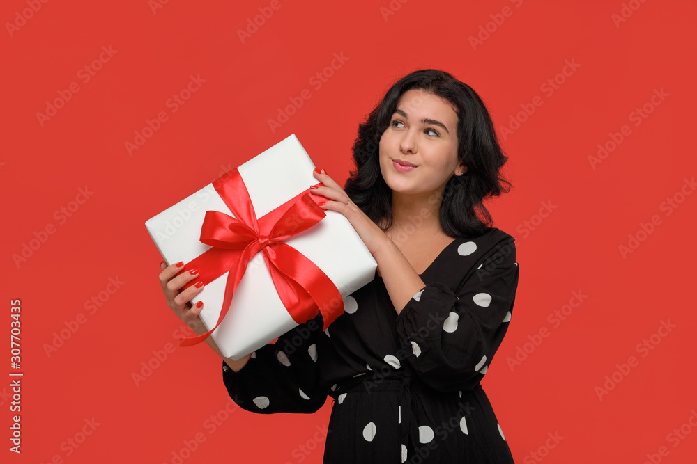 Brunette woman in a black dress smiling holding Christmas giftbox of white color with red ribbon. Brunette woman in a black dress smiling holding Christmas giftbox of white color with red ribbon.