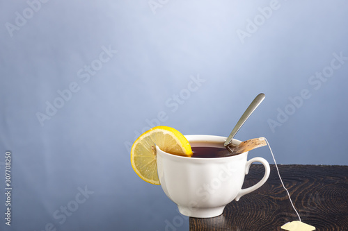 white mug with tea and bag, lemon and a spoon inside on the edge of the table on a gray background
