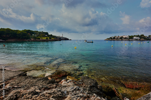 Transparent and colorful waters in beautiful bay of the south of Mallorca. In the background lighthouse and rocks in the foreground. Cloudy sky.
