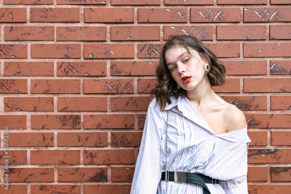 Waist up portrait of stylish brunette girl with red lips and wavy brunette hair dressed in white striped shirt with naked shoulder isolated over red brick wall
