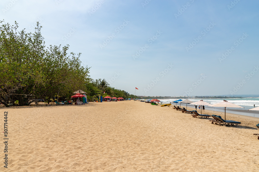 Fototapeta premium KUTA / INDONESIA - OCTOBER 25, 2019: Wide sand beach with tourists, umbrellas and beds.