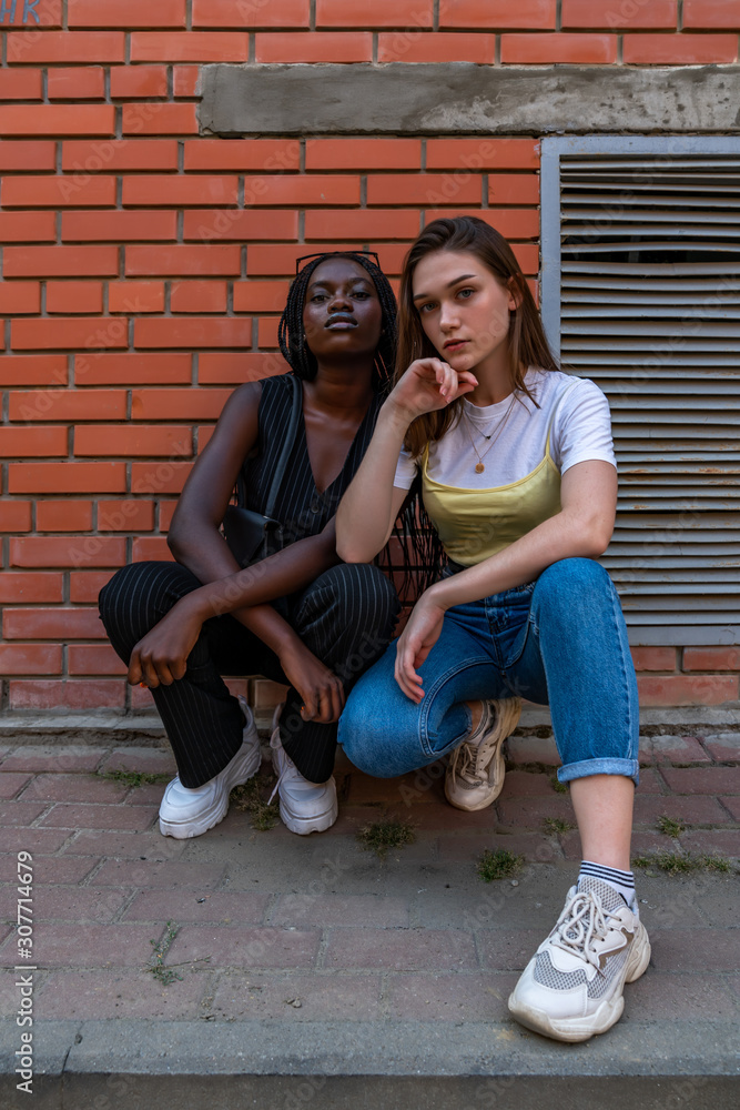 Multi ethnic stylish portrait of young girls in casual clothes that crouched and looking at the camera isolaed over red brick wall