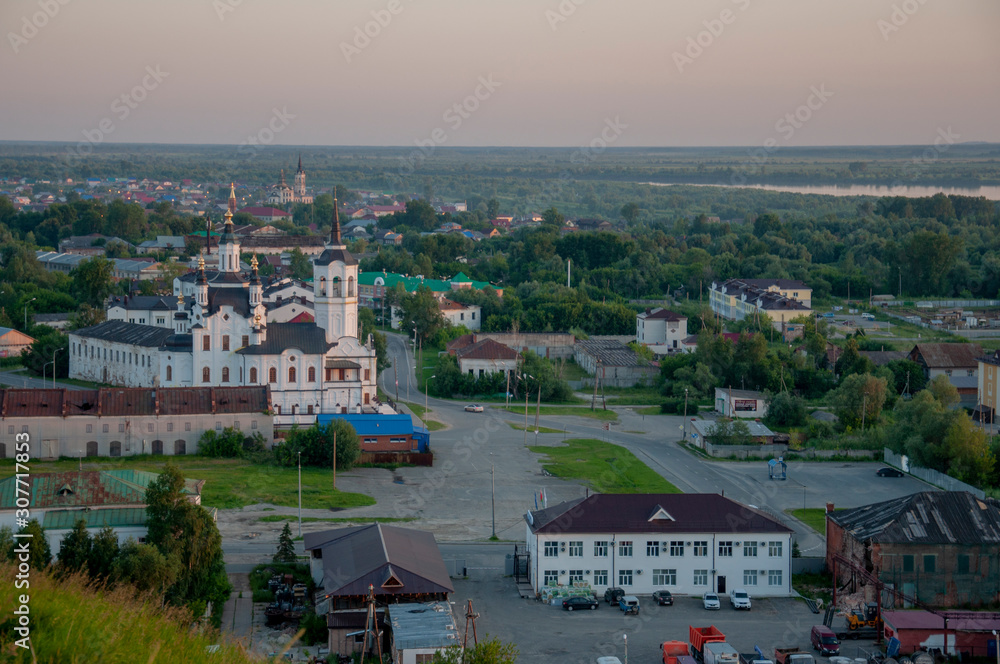 Fototapeta premium Small town with tiny houses near the river and green forests and fields. Trees and their shadows on the grass. Sundown and sunrises. View from high on the landscape