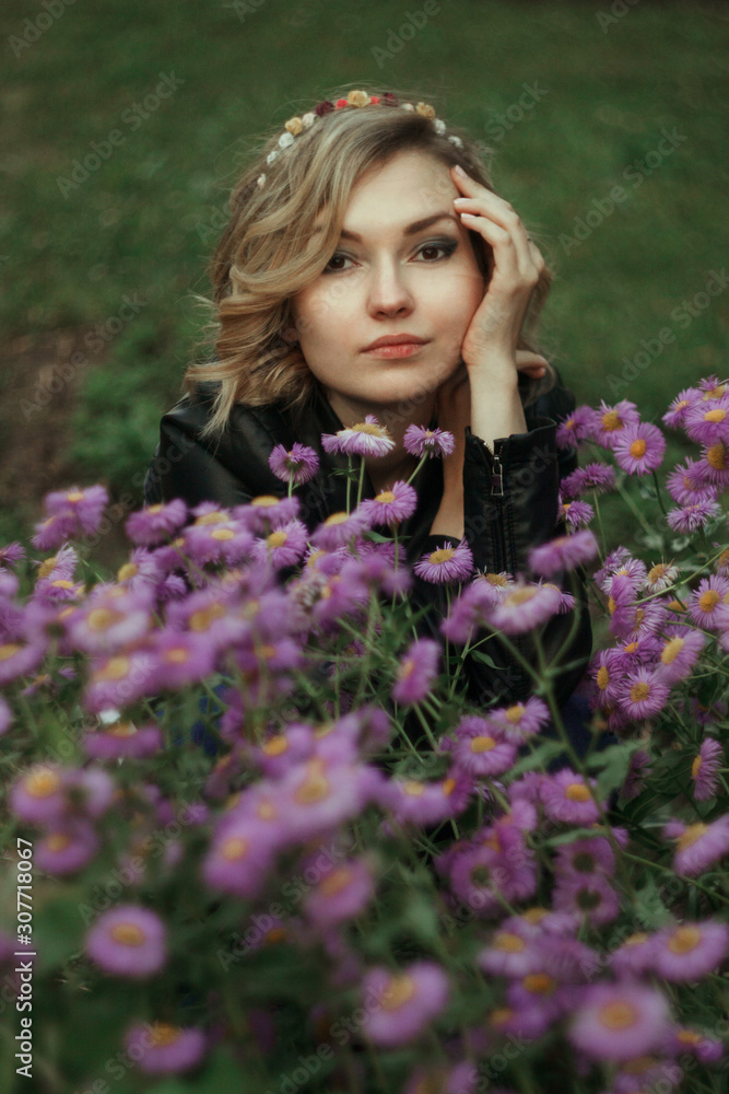 Portrait of a young woman with curly white hair in a black sweater and in a wreath on the street in a summer park on a background of green trees. Purple flowers in the foreground Portrait of a young woman with curly white hair in a black sweater and in a wreath on the street in a summer park on a background of green trees. Purple flowers in the foreground