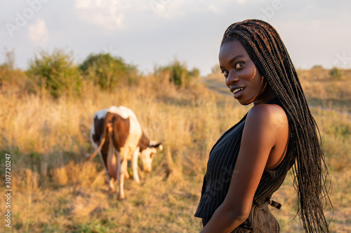 Waist up portrait of the toung african girl in black clothes stands among the field and looking at the camera over her shoulder, cow graze on the background © Vasya