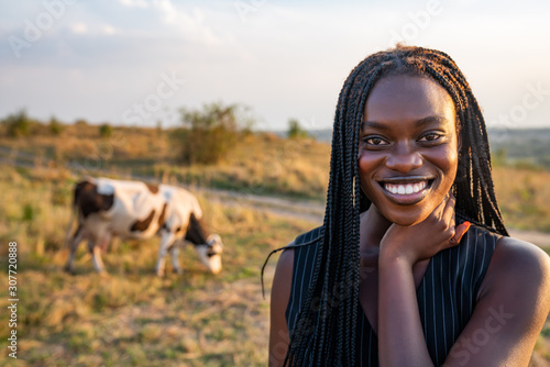 Close up portrait of the young african girl in black vest among the field, cow graze on the background photo