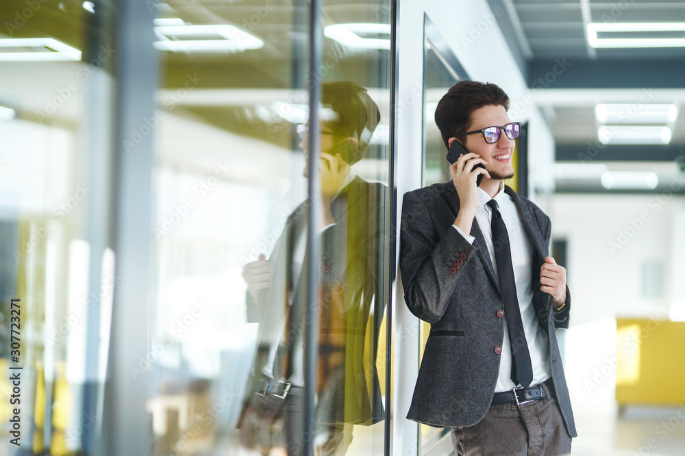 Young businessman in glasses talking on cell phone standing in the modern office. Stylish man in a suit with folder standing and talking on telephone. Communication concept. Young businessman in glasses talking on cell phone standing in the modern office. Stylish man in a suit with folder standing and talking on telephone. Communication concept.