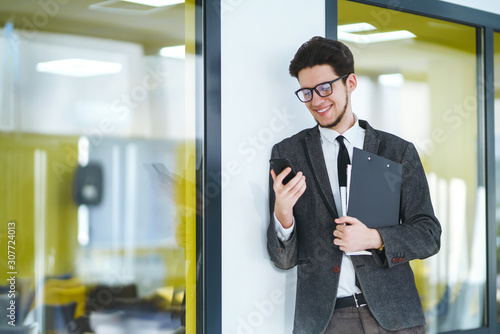 Young office worker in glasses using mobile smart phone. Businessman holds telephone in hand. Stylish man in a suit with phone in hands prints a message on the phone. Communication concept.