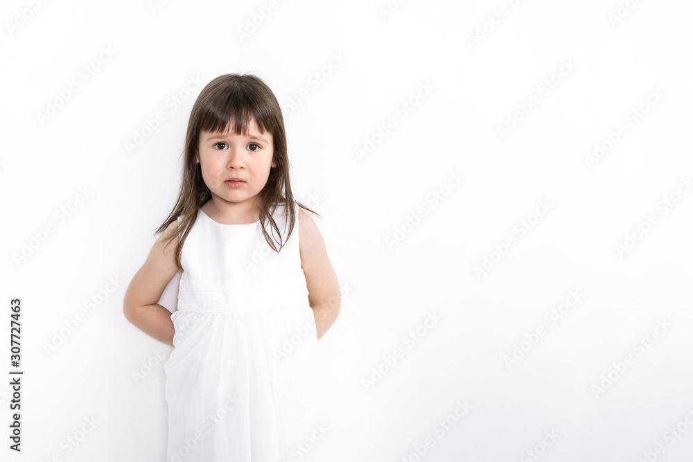 A sad and frightened girl is standing on a white background. A confused child is standing against the wall. A sad and frightened girl is standing on a white background. A confused child is standing against the wall.