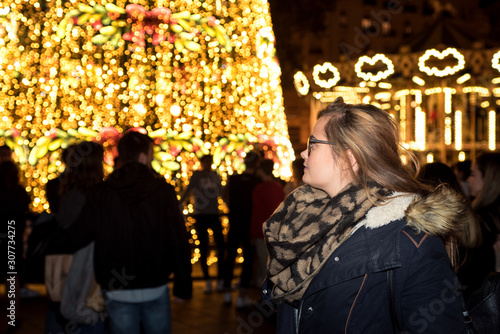 Blonde girl with glasses  looking at the Christmas lights of the city square. View from behind