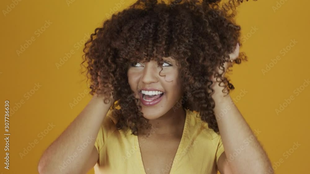 Happy laughing American African woman with her cyrly hair in yellow background. Laughing curly woman in sweater touching her hair and looking at the camera. Happy laughing American African woman with her cyrly hair in yellow background. Laughing curly woman in sweater touching her hair and looking at the camera.
