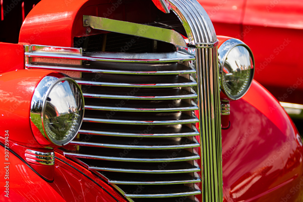Old vintage American red pickup car front half side view close up, chrome headlights light lamp and grille, with open hood during an outdoor show