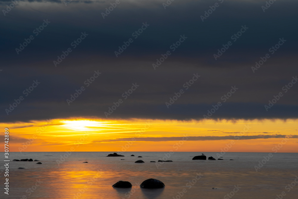 Vibrant colored winter sunset reflecting in ocean with endless horizon and deep blue ocean, silhouette of boulders laying in the foreground in shallow water at island of Gotland, Sweden