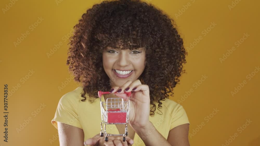 Girl with small shopping card. Smiling and dancing curly hair woman in shopping concept. Young woman with a miniature cart. E-commerce and business. Shopping car. Woman shopper. Yellow background. 4K. Girl with small shopping card. Smiling and dancing curly hair woman in shopping concept. Young woman with a miniature cart. E-commerce and business. Shopping car. Woman shopper. Yellow background. 4K.