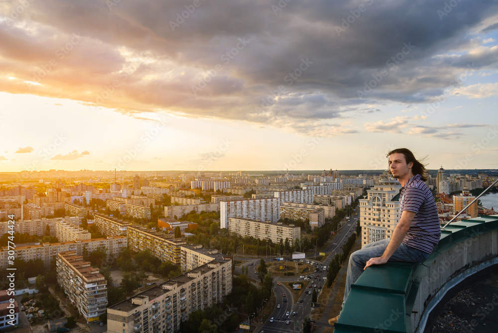 Young and brave man sitting on the edge of the roof and looking far away at the city, sunset landscape