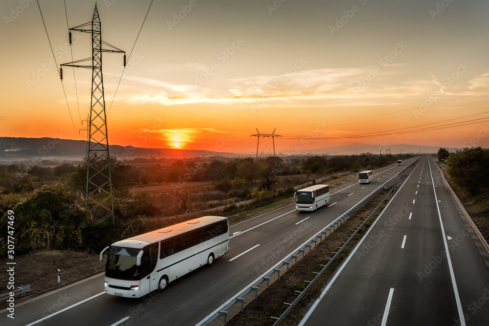Caravan or convoy of Four buses in line traveling on a country highway under amazing orange   sunset sky. Highway transportation with white buses