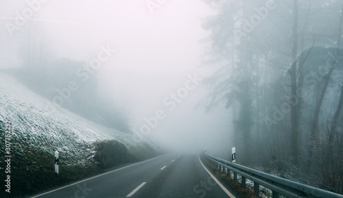 Landscape of lonely paved road running away among slope in slight snow and coniferous trees in thick fog