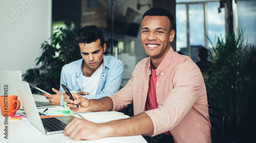 Portrait of cheerful African American male employee with cellphone gadget sitting at office table and smiling at camera enjoying work occupation, diverse colleagues braistorming and cooperating
