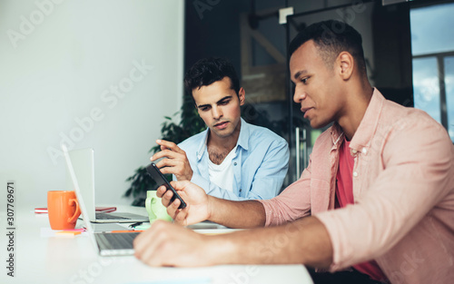 Two millennials reading received email message during mobile chatting in office interior connecting to 4g wireless for surfing web internet, diverse analysts browsing useful data information