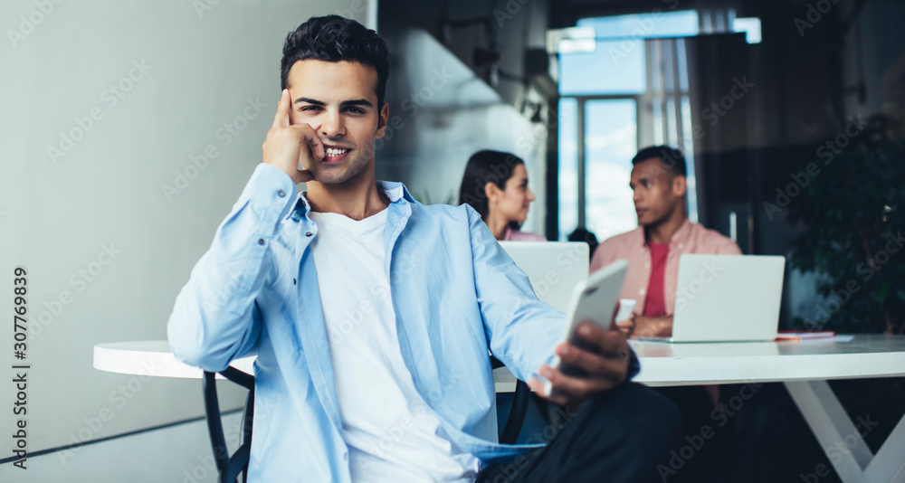 Obraz premium Portrait of happy millennial man holding cellphone device for communication in hand and smiling at camera during work day in office space, Spanish generation Y enjoying job experience indoors