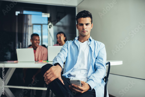 Portrait of handsome millennial man holding cellphone device for communication in hand and looking at camera during work day in office space, Spanish male generation Y with smartphone gadget