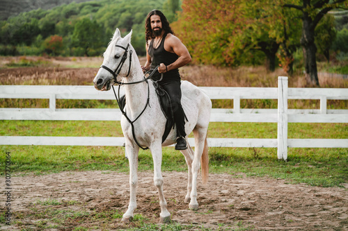 Rider on gray arabian horse in the field. Handsome bearded man riding horse at farm. Beautiful horse with man rider trotting on autumnal field. Equestrian and animal love concept.