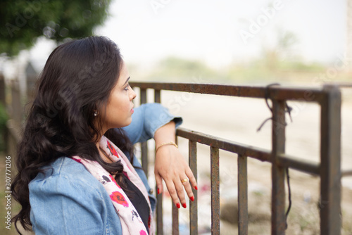 An young and attractive Indian Bengali woman in blue western jacket is standing holding a railing in green countryside background. Indian lifestyle.