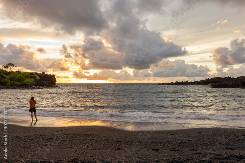 A woman watches the sunrise at Honokalani black sand beach