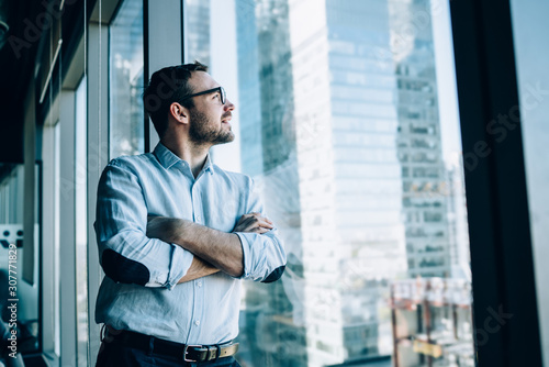 Contemplative male entrepreneur with crossed hands standing near office window view and feeling pondering during work day in company, Caucasian pensive corporate boss thoughtful looking away photo