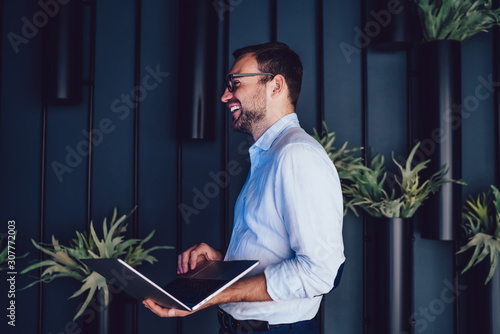 Cheerful male executive manager in formal clothing holding laptop computer in hands and laughing during corporate messaging with colleagues, happy boss using netbook in modern workspace interior photo