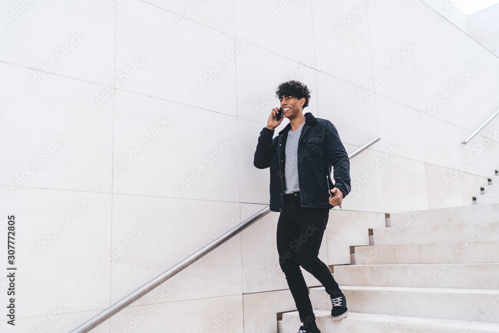 Fototapeta premium Trendy cheerful African American man speaking on phone while walking on urban staircase on street
