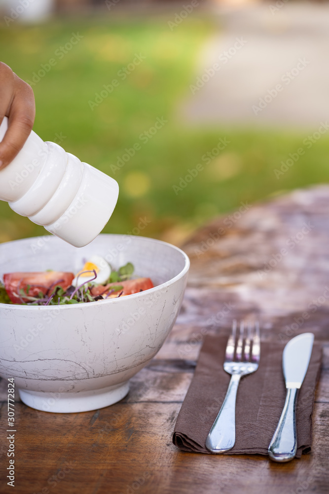 Hand with a white pepper shaker seasoning a fresh salad bowl mixed green  leaves, eggs, black olives and tomato on a wooden table with cutlery.  Blurred nature background. Vertical photography Stock Photo