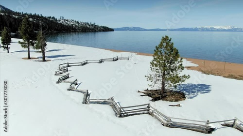 Aerial: Flying over pine tree surrounded by a fence and snow on the ground overlooking a calm & tranquil Lake Tahoe in the winter time. In the background are mountains covered in snow. Nevada, USA